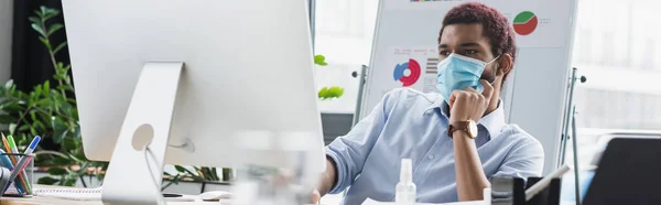 Африканский американский бизнесмен в медицинской маске смотрит на монитор компьютера в офисе, баннер — стоковое фото