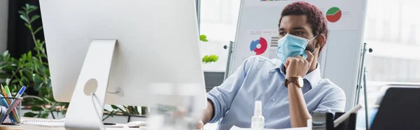 Hombre de negocios afroamericano en máscara médica mirando el monitor de computadora en la oficina, pancarta - foto de stock