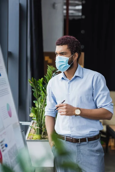 Homme d'affaires afro-américain en masque médical regardant le tableau à feuilles mobiles au bureau — Photo de stock
