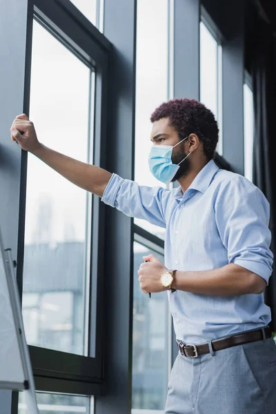 Jeune homme d'affaires afro-américain en masque médical debout près de la fenêtre et tableau à feuilles mobiles — Photo de stock