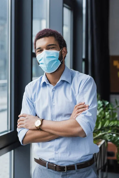 Африканский американский бизнесмен в медицинской маске стоит со скрещенными руками в кабинете — стоковое фото