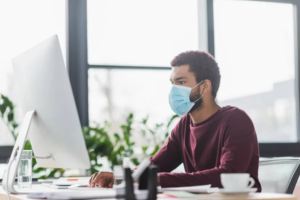 Empresário afro-americano em máscara médica usando computador perto de café e água no escritório — Fotografia de Stock