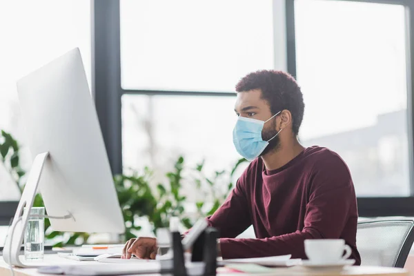 Uomo d'affari afroamericano in maschera medica utilizzando il computer vicino a caffè e acqua in ufficio — Foto stock