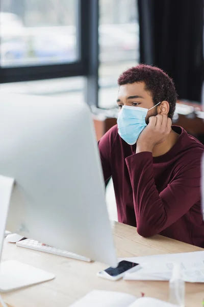 Hombre de negocios afroamericano en máscara médica mirando el monitor de la computadora cerca de teléfono inteligente y desinfectante de manos - foto de stock