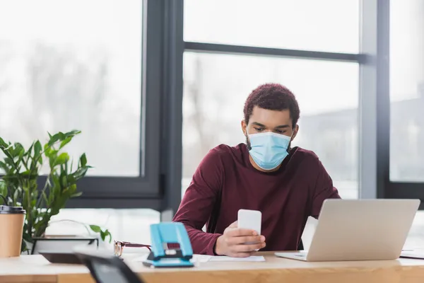 Uomo d'affari afroamericano in maschera protettiva utilizzando il telefono cellulare vicino laptop e caffè per andare in ufficio — Foto stock