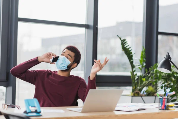 Uomo d'affari afroamericano in maschera medica che parla su smartphone vicino a documenti e laptop in ufficio — Foto stock