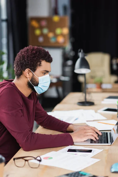 Vista lateral de un joven hombre de negocios afroamericano en máscara médica usando un portátil cerca de papeles y anteojos en la oficina - foto de stock