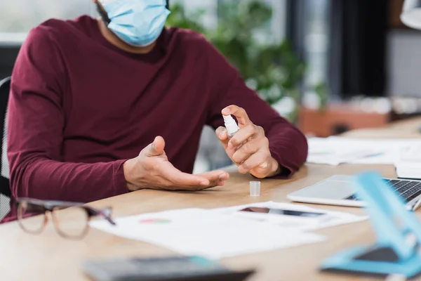 Vista recortada del hombre de negocios afroamericano en máscara médica usando desinfectante de manos cerca de dispositivos en la oficina - foto de stock