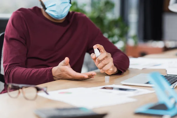 Cropped view of african american businessman in medical mask using hand sanitizer near devices in office — Stock Photo