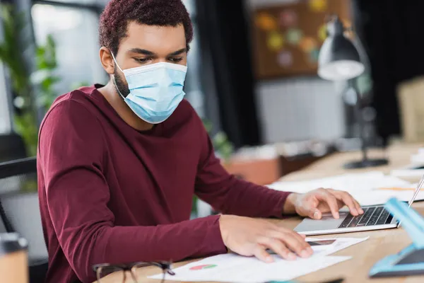 Африканский американский бизнесмен в медицинской маске работает с устройствами и бумагами в офисе — стоковое фото