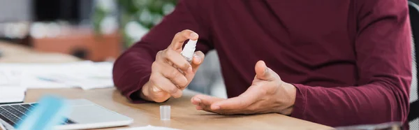 Vista ritagliata di uomo d'affari afro-americano che utilizza disinfettante per le mani vicino al laptop in ufficio, banner — Foto stock