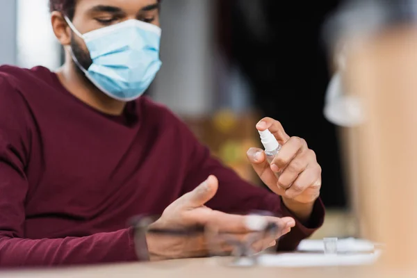 Empresario afroamericano borroso en máscara médica usando desinfectante de manos en la oficina - foto de stock