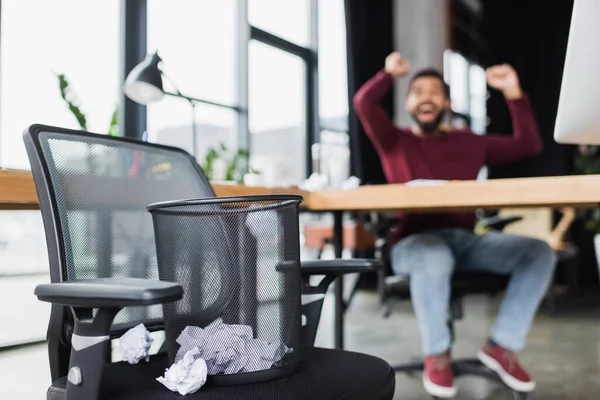 Lata de lixo com papel amassado na cadeira perto de homem de negócios afro-americano borrado no escritório — Fotografia de Stock