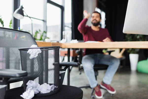 Zerknülltes Papier neben Mülleimer und verschwommener afrikanischer Geschäftsmann im Amt — Stockfoto