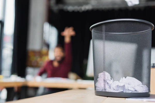 Trash can with crumpled paper near blurred african american businessman in office — Stock Photo