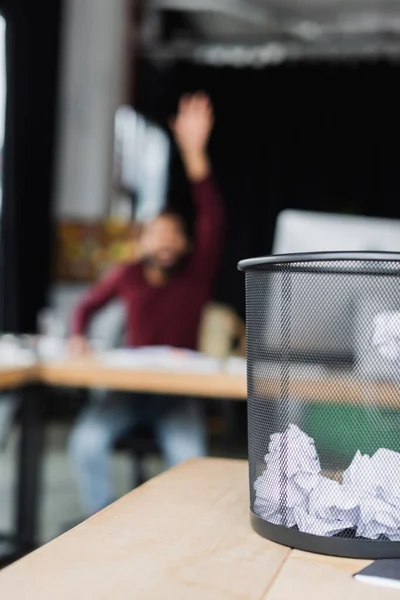 Trash can with crumpled paper on table near blurred african american businessman in office — Stock Photo