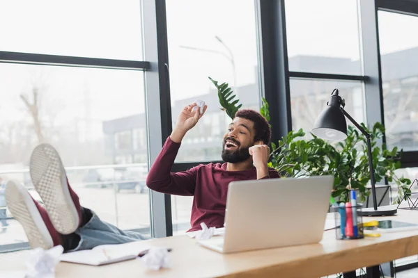 Positive african american businessman holding crumpled paper near laptop in office — Stock Photo