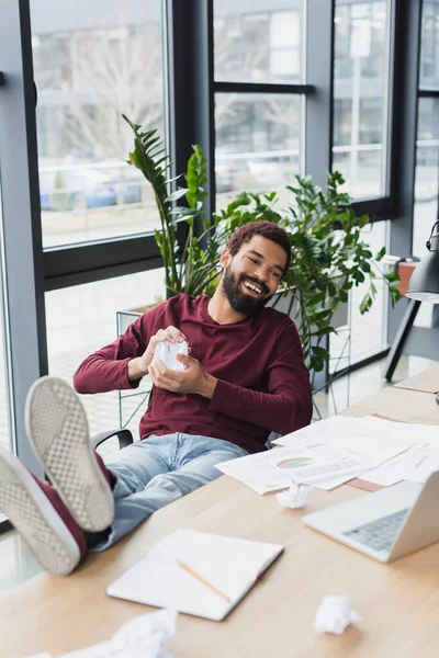 Sorridente uomo d'affari africano americano in possesso di carta accartocciata vicino al computer portatile in ufficio — Foto stock