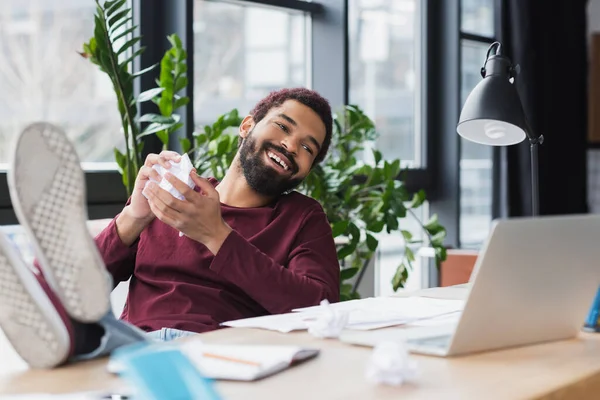 Happy african american businessman holding crumpled paper near blurred laptop and lamp in office — Stock Photo