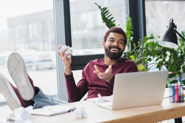 Empresário afro-americano positivo segurando papel amassado durante videochamada no laptop no escritório — Fotografia de Stock