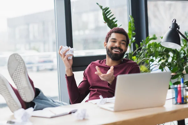 Un homme d'affaires afro-américain positif tenant du papier froissé pendant un appel vidéo sur un ordinateur portable au bureau — Photo de stock