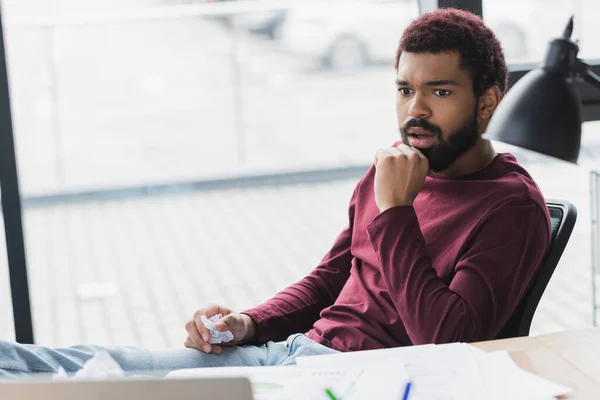 Hombre de negocios afroamericano preocupado con papel arrugado mirando a la computadora portátil en la oficina - foto de stock