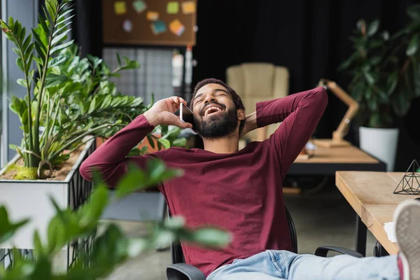 Fröhlicher afrikanisch-amerikanischer Geschäftsmann spricht auf Smartphone in der Nähe von Pflanzen im Büro — Stockfoto