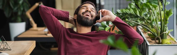 Alegre hombre de negocios afroamericano hablando por celular cerca de plantas en la oficina, pancarta - foto de stock