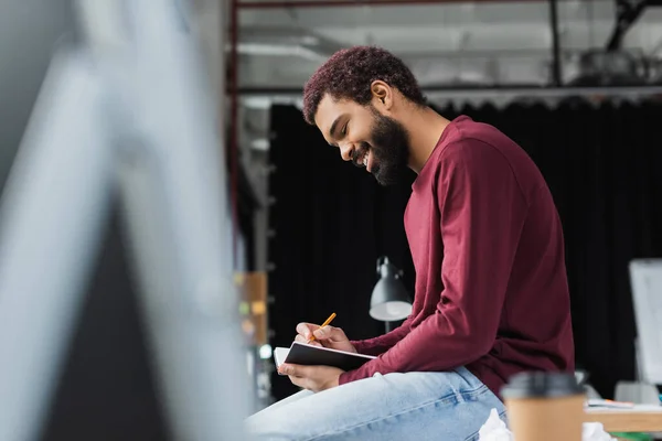 Vista lateral del sonriente hombre de negocios afroamericano escribiendo en un cuaderno cerca de café borroso para ir en la oficina - foto de stock