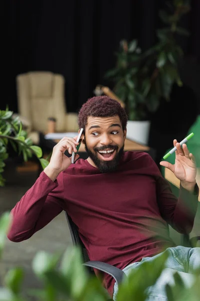 Emocionado hombre de negocios afroamericano sosteniendo marcador mientras hablaba en el teléfono inteligente en la oficina - foto de stock