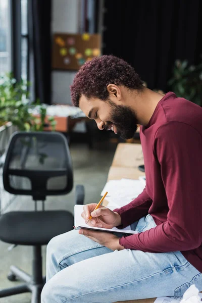 Seitenansicht eines lächelnden afrikanisch-amerikanischen Geschäftsmannes, der im Büro auf Notizbuch schreibt — Stockfoto