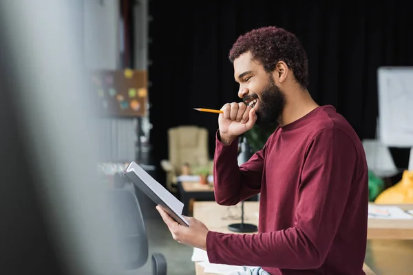Vista lateral do empresário afro-americano positivo segurando lápis e caderno no escritório — Fotografia de Stock