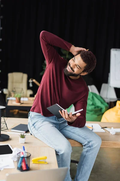 Щасливий афроамериканець у звичайному одязі, який тримає записник біля робочого столу на посаді. — стокове фото
