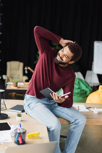 Happy african american businessman in casual clothes holding notebook near working table in office — Stock Photo