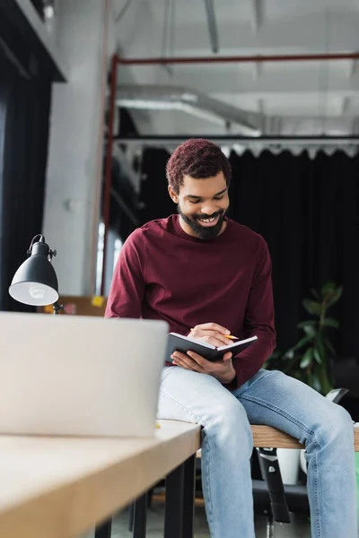 Positivo uomo d'affari afro-americano che scrive sul taccuino mentre siede sul tavolo vicino a un computer portatile offuscato — Foto stock