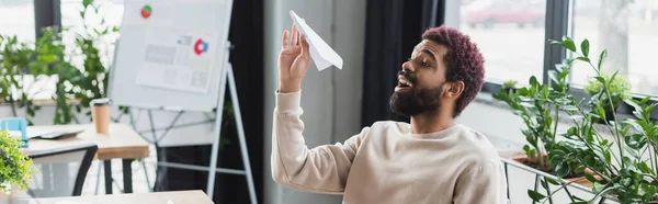 Cheerful african american businessman holding paper plane in office, banner — Stock Photo