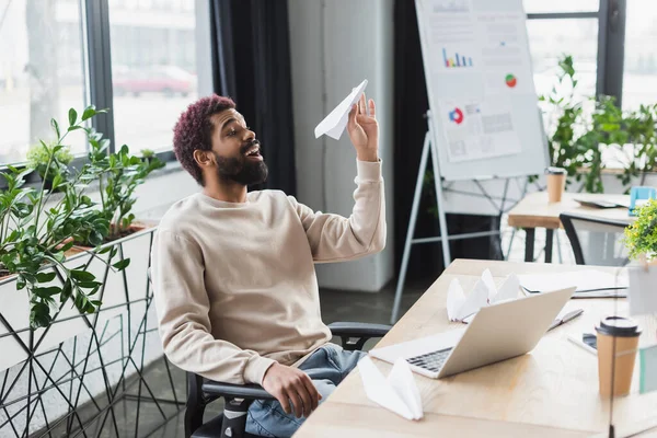 Joven empresario afroamericano sosteniendo avión de papel cerca de la computadora portátil y café para ir en la oficina - foto de stock