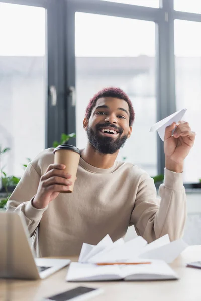 Empresário afro-americano positivo segurando café para ir e avião de papel perto de laptop no escritório — Fotografia de Stock