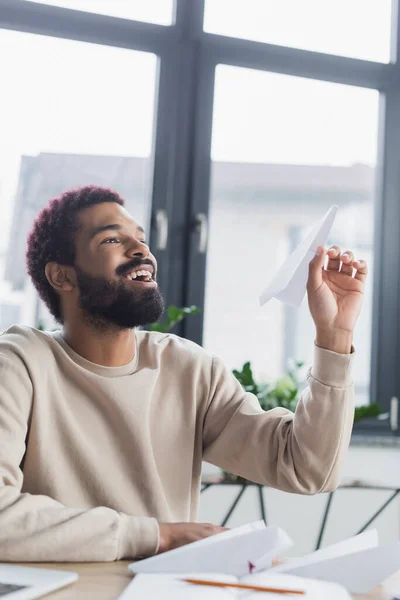 Sonriente hombre de negocios afroamericano sosteniendo avión de papel en la oficina - foto de stock