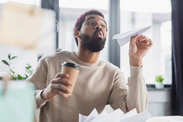 African american businessman in casual clothes holding paper plane and coffee to go in office — Stock Photo