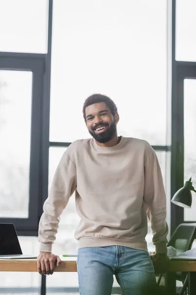 Sorrindo empresário afro-americano olhando para a câmera perto da mesa no escritório — Fotografia de Stock