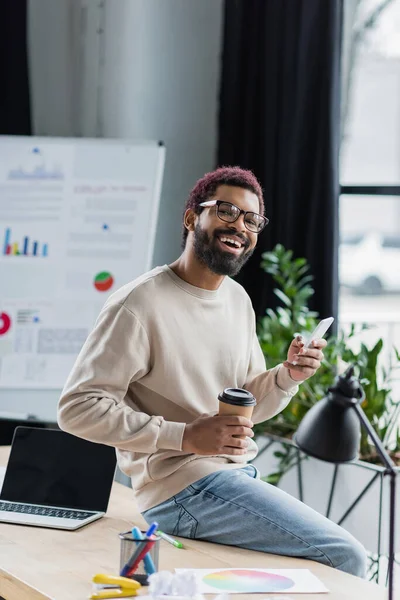 Sonriente hombre de negocios afroamericano sosteniendo teléfono inteligente y café para ir en la oficina - foto de stock