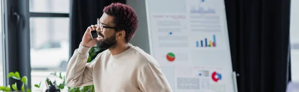 Sonriente hombre de negocios afroamericano en gafas que hablan en el teléfono inteligente en la oficina, pancarta - foto de stock