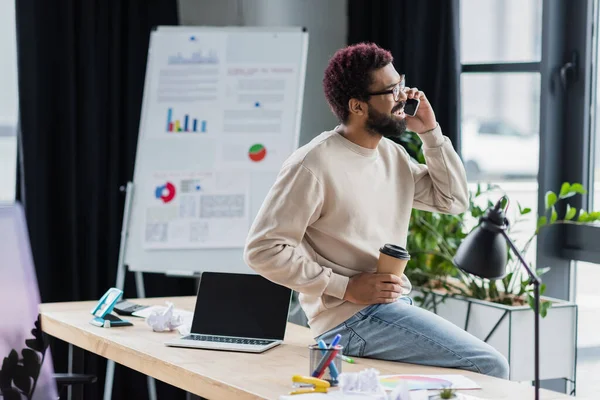 Positivo uomo d'affari afroamericano in occhiali che parla su smartphone e tiene in mano il drink da asporto vicino al laptop in ufficio — Foto stock