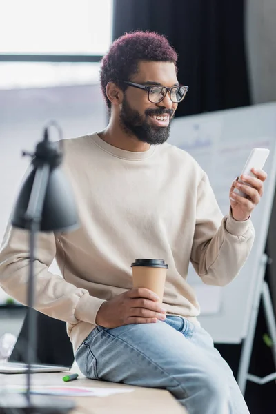 Felice uomo d'affari afro-americano utilizzando smartphone e tenendo in carica la tazza di carta — Foto stock