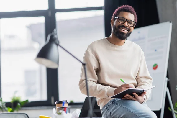 Cheerful african american businessman writing on notebook in office — Stock Photo