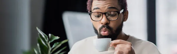 African american businessman in eyeglasses blowing at cup of coffee in office, banner — Stock Photo