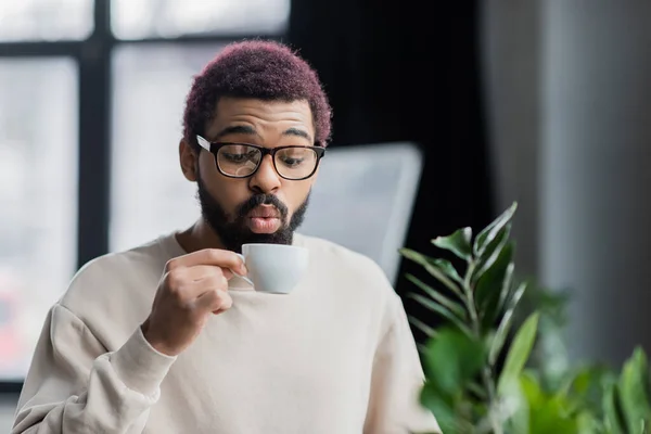 Молодой африканский бизнесмен в очках, дующий за чашкой кофе в офисе — стоковое фото