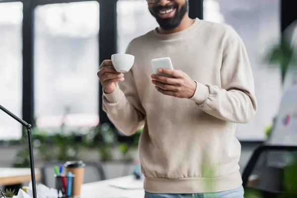 Ausgeschnittene Ansicht von Smartphone und Tasse in den Händen eines verschwommenen afrikanisch-amerikanischen Geschäftsmannes im Büro — Stockfoto