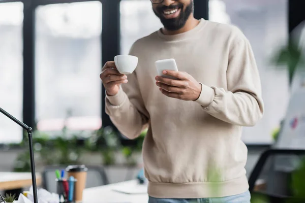 Vue recadrée du smartphone et de la tasse entre les mains d'un homme d'affaires afro-américain flou au bureau — Photo de stock